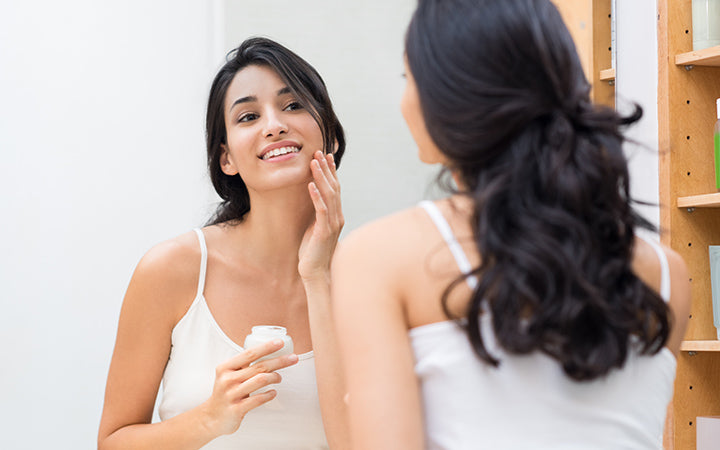 Vrouw die zorgt voor haar mooie huid op het gezicht dat in de buurt van de spiegel staat en vochtinbrengende crème op haar gezicht aanbrengt